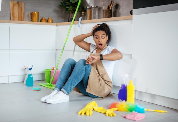 Sorprendida dama negra milenaria se sienta en el piso con suministros de limpieza de trapeador con la boca abierta mira el reloj