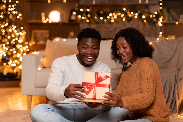 Sorprendida feliz joven pareja negra abre caja con regalo en el acogedor salón interior con árbol de Navidad con luces