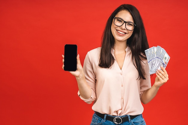 Sorprendida, feliz y sonriente mujer morena mostrando la pantalla de un smartphone en blanco y señalándola mientras miraba la cámara con una sonrisa aislada sobre el fondo rojo Sosteniendo billetes de dinero