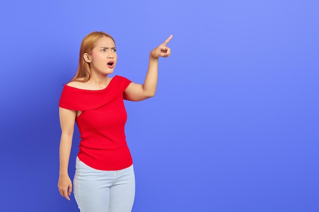 Sorprendida hermosa mujer asiática señalando con el dedo el espacio de copia aislado en el fondo púrpura