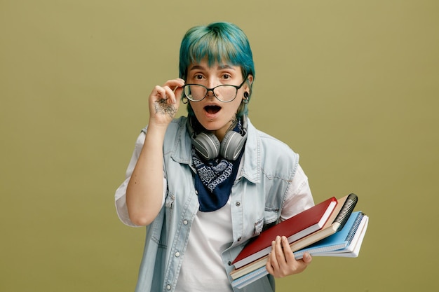 Sorprendida joven estudiante con anteojos bandana y audífonos alrededor del cuello sosteniendo blocs de notas agarrando anteojos mirando la cámara desde detrás de anteojos aislados en un fondo verde oliva