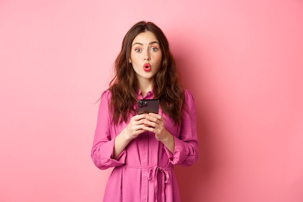 sorprendida joven jadeando y diciendo wow, mirando impresionado a la cámara después de leer noticias en línea en el teléfono móvil, de pie con el teléfono inteligente contra la pared rosa. Copia espacio