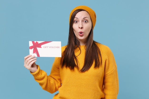 Sorprendida joven morena con suéter amarillo y sombrero posando aislada en un retrato de estudio de fondo azul. Gente emociones sinceras concepto de estilo de vida. Simulacros de espacio de copia. Mantenga el certificado de regalo.