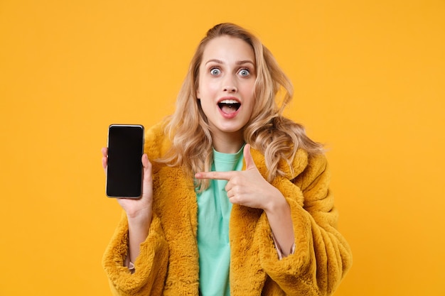 Sorprendida joven mujer rubia con abrigo de piel amarillo posando aislada de fondo naranja. Concepto de estilo de vida de las personas. Simulacros de espacio de copia. Señalar con el dedo índice en el teléfono móvil con pantalla vacía en blanco.