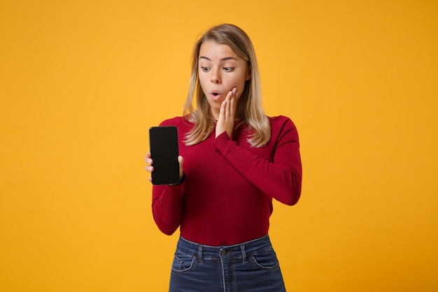 Sorprendida joven mujer rubia con ropa informal posando aislada en un fondo amarillo anaranjado. Concepto de estilo de vida de las personas. Simulacros de espacio de copia. Sostenga el teléfono móvil con la pantalla vacía en blanco y ponga la mano en la mejilla.