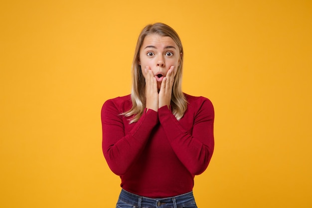 Sorprendida joven mujer rubia con ropa informal posando aislada en un retrato de estudio de fondo naranja amarillo. Concepto de estilo de vida de las personas. Simulacros de espacio de copia. Poner las manos en las mejillas, manteniendo la boca abierta.