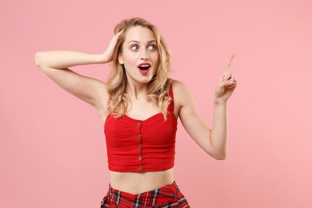 Sorprendida joven mujer rubia con ropa sexy roja posando aislada en un fondo rosa pastel en el estudio. Concepto de estilo de vida de las personas. Simulacros de espacio de copia. Poner la mano en la cabeza apuntando con el dedo índice a un lado.