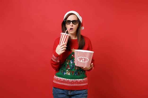 Sorprendida joven Santa en gafas imax 3d viendo una película, sosteniendo una taza de refresco de palomitas de maíz aislado sobre fondo de pared roja. Feliz año nuevo 2019 celebración concepto de fiesta navideña. Simulacros de espacio de copia.