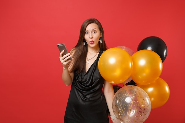 Sorprendida joven sorprendida en pequeño vestido negro sosteniendo globos de aire con teléfono móvil mientras celebra aislado sobre fondo rojo. Día de la mujer feliz año nuevo, concepto de fiesta de vacaciones de maqueta de cumpleaños.