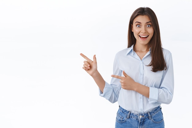 Foto sorprendida joven trabajadora exitosa en blusa azul se ve impresionada al ver algo inesperado e impresionante, señalando con el dedo a la izquierda y compartiendo una gran noticia con usted, pared blanca