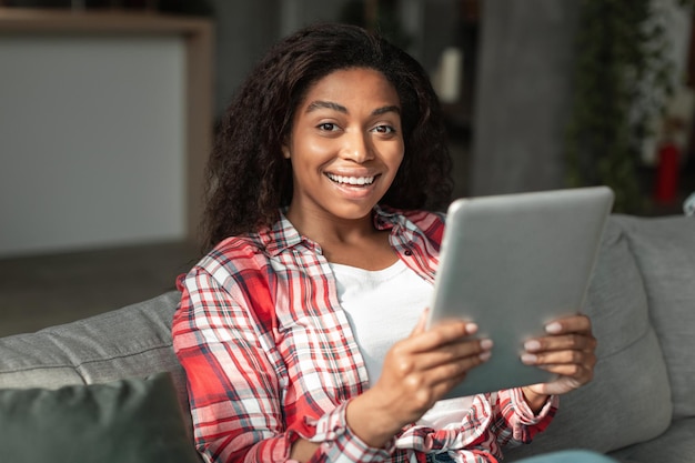 Sorprendida mujer afroamericana milenaria feliz escribiendo en la tableta en el sofá en el interior de la sala de estar