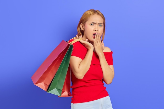 Sorprendida mujer asiática joven sosteniendo bolsas de compras con la boca abierta y mirando a la cámara aislada sobre fondo púrpura