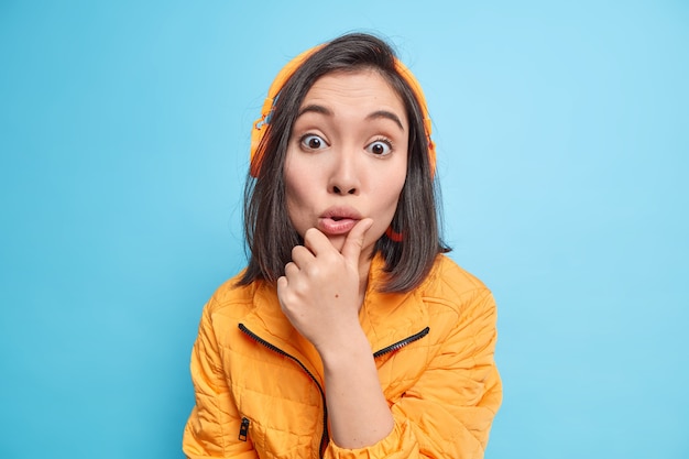 sorprendida mujer asiática mira con asombro usa auriculares estéreo en los oídos escucha música de la lista de reproducción vestida con una chaqueta naranja aislada sobre una pared azul. Emociones humanas