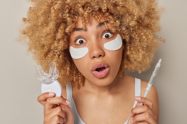 Sorprendida mujer joven con cabello rubio rizado sostiene un expansor de boca de plástico y gel en jeringa aplica parches de belleza debajo de los ojos para eliminar las arrugas mantiene la mandíbula caída aislada sobre la pared gris