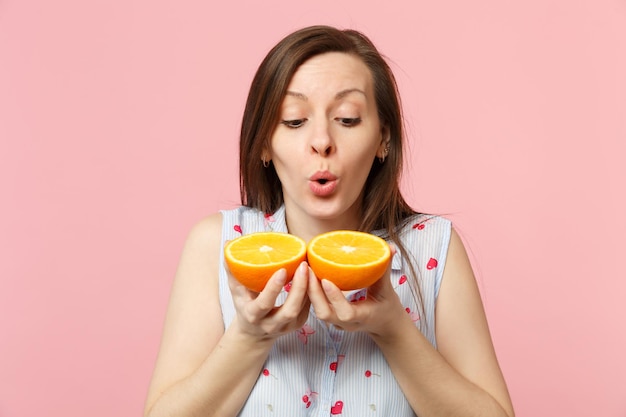 Sorprendida mujer joven con ropa de verano sosteniendo en las manos dos mitades de fruta fresca de naranja madura aislada en un fondo rosa pastel. El estilo de vida vívido de la gente relaja el concepto de vacaciones. Simulacros de espacio de copia.