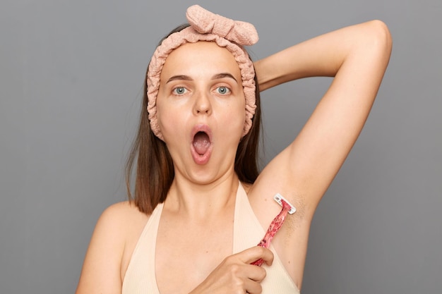 Sorprendida mujer joven sorprendida con una banda para el cabello usando una maquinilla de afeitar para quitarse el pelo de la axila hablando de su belleza e higiene posando aislada sobre fondo gris