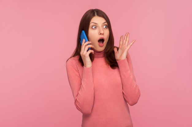 Sorprendida mujer maravillada con cabello castaño en suéter rosa hablando por teléfono escuchando información con expresión sorprendida increíble tiro de estudio interior aislado sobre fondo rosa