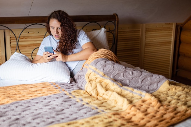 Sorprendida mujer morena rizada acostada en la cama bajo una manta con la boca abierta sosteniendo el teléfono en las manos y mirando el teléfono usando pijamas de casa durante el día