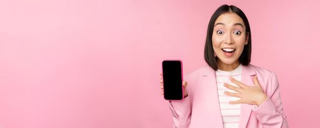 Sorprendida mujer de negocios asiática entusiasta que muestra la interfaz de la aplicación del teléfono inteligente de la pantalla del teléfono móvil de pie contra el fondo rosa