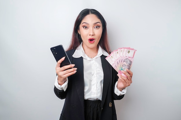 Sorprendida mujer de negocios asiática con traje negro sosteniendo su teléfono inteligente y dinero en rupia indonesia aislada de fondo blanco
