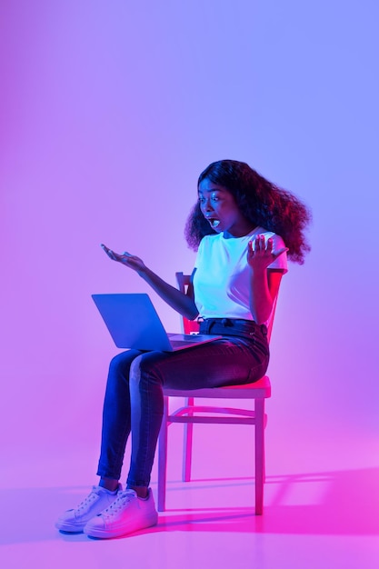 Sorprendida mujer negra milenaria usando una laptop abriendo la boca sorprendida sentada en una silla con luz de neón