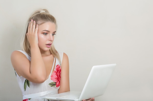 Sorprendida mujer rubia atractiva mirando a la computadora portátil, posando sobre el fondo gris, espacio para texto