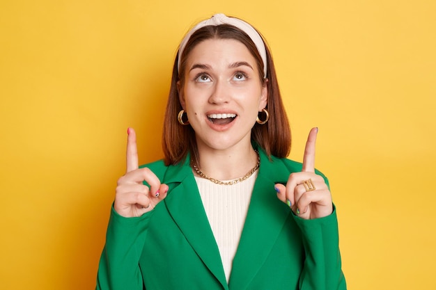 Sorprendida mujer sorprendida con chaqueta verde posando aislada sobre fondo amarillo apuntando hacia el espacio de copia para el área de publicidad de promoción