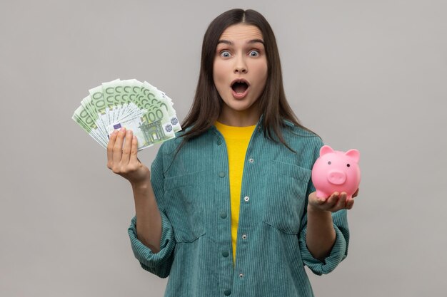 Sorprendida mujer sorprendida sosteniendo una gran suma de billetes en euros y una inversión rentable de la alcancía