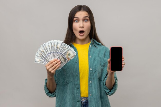 Sorprendida mujer sorprendida sosteniendo en las manos un teléfono inteligente con pantalla en blanco y billetes de dólares