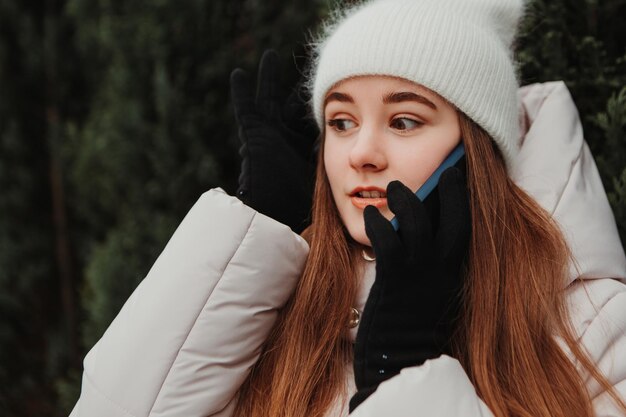 Sorprendida mujer sorprendida vestida con una chaqueta abrigada, parada cerca del árbol de Navidad, hablando por teléfono gesticulando