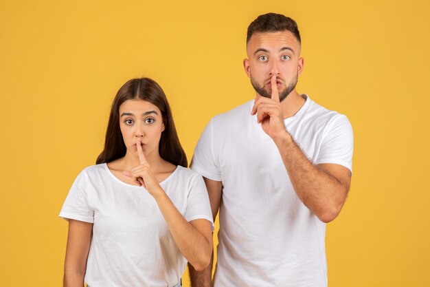 Sorprendida pareja joven con camisetas blancas con los dedos en los labios haciendo un gesto de silencio