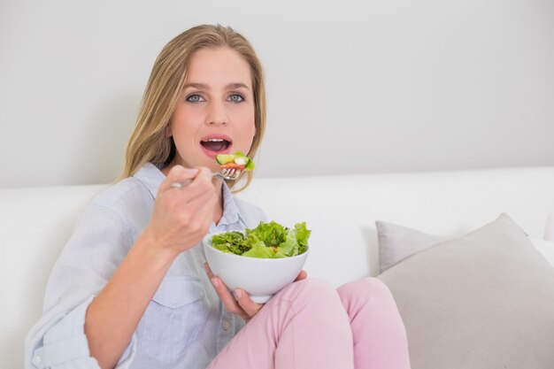 Sorprendida rubia casual sentada en el sofá comiendo ensalada