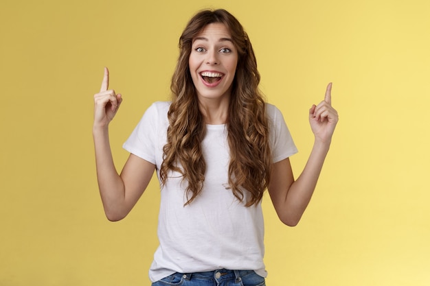 Sorprendido alegre celebrando a la chica animada discutir cerca de un evento impresionante apuntando hacia arriba con los dedos índices sonriendo ampliamente fascinado mirar temtation admirar gran concierto stand fondo amarillo impresionado.