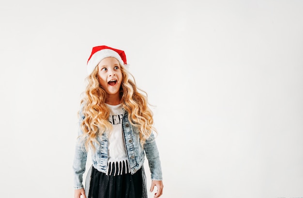 Sorprendido chica de cabello rizado entre santa sombrero y chaqueta de mezclilla y falda de tutú negro sobre blanco aislado