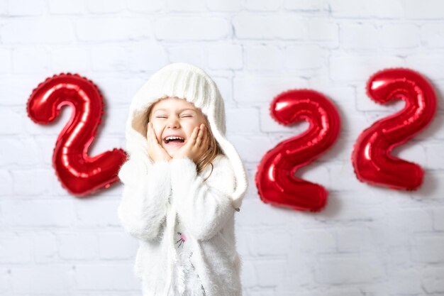 Sorprendido, feliz, emocionado, sonriente, positivo, niño, niña, en, tejido, sombrero, encima, blanco, descanso, pared, plano de fondo, con, cifras, rojo, globos, año nuevo, números, 2022