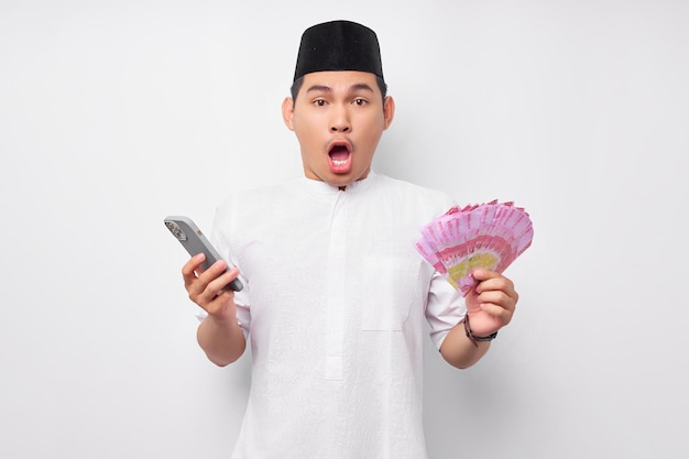 Sorprendido guapo musulmán asiático sosteniendo teléfono móvil y billetes de rupias de dinero en efectivo aislado sobre fondo blanco Concepto de estilo de vida islámico religioso de la gente