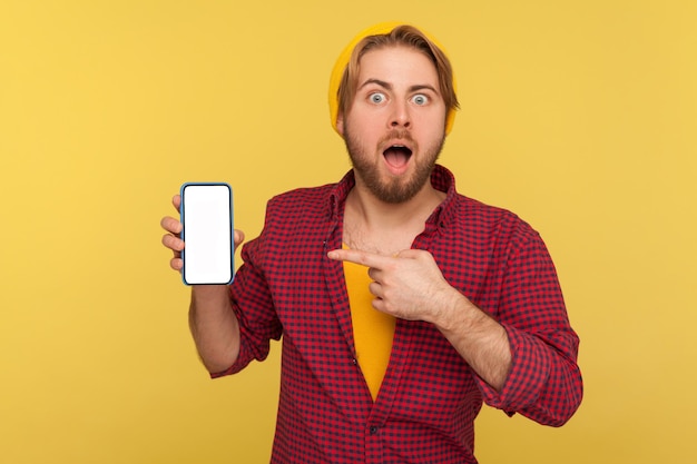 Sorprendido hipster de moda con sombrero de beanie y camisa a cuadros apuntando a la pantalla vacía del teléfono móvil, mirando a la cámara con asombro, mostrando la maqueta en blanco del teléfono móvil para publicidad. tiro del estudio aislado