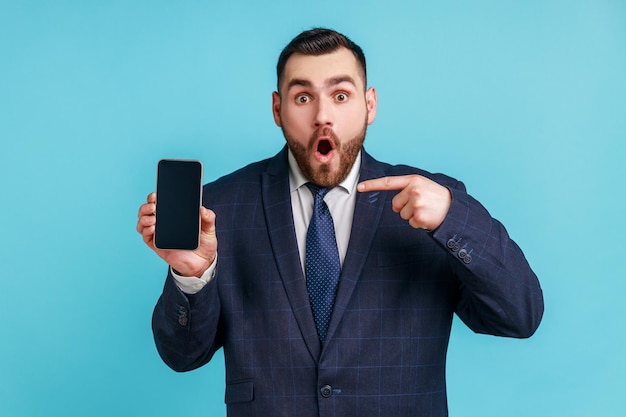 Sorprendido hombre barbudo con traje de estilo oficial apuntando con el dedo al smartphone con pantalla blanca vacía, impactado con las nuevas características del teléfono. Disparo de estudio interior aislado sobre fondo azul.