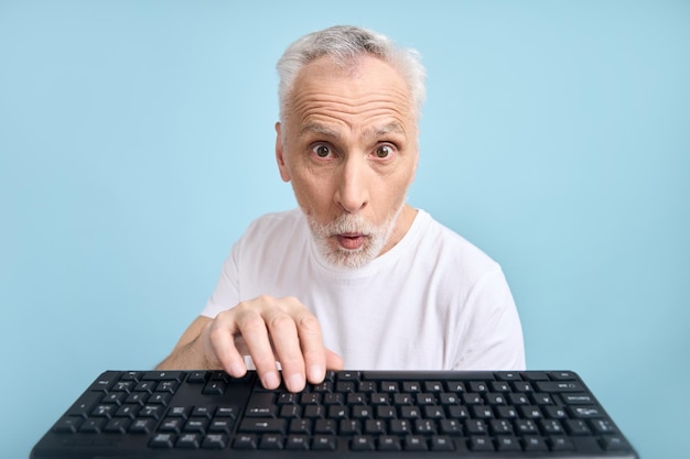 Sorprendido hombre mayor de unos 60 años sosteniendo la mano en el teclado de la computadora expresando sorpresa poniendo los ojos en blanco y posando con la boca abierta sobre un fondo azul aislado Personas Tecnología Laptop POV