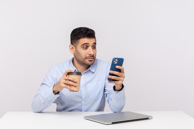 Sorprendido hombre de negocios sentado en el lugar de trabajo con café en la mano y revisando el mensaje leyendo el correo electrónico en el teléfono móvil con una expresión sorprendida en un estudio interior aislado en fondo blanco