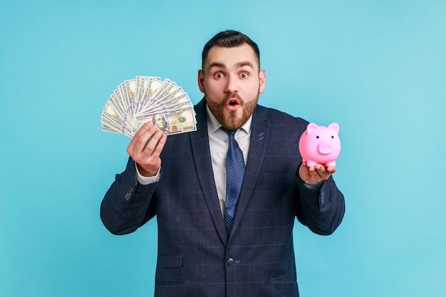 Sorprendido hombre de negocios con traje de estilo oficial mirando a la cámara con una expresión impactante, sosteniendo alcancías y billetes en dólares en las manos. Disparo de estudio interior aislado sobre fondo azul.