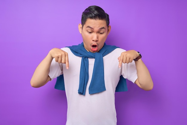 Sorprendido joven asiático con camiseta blanca apuntando con el dedo hacia abajo mostrando el espacio de copia aislado sobre el concepto de estilo de vida de la gente de fondo púrpura