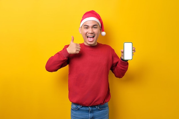 Sorprendido joven asiático con sombrero de Papá Noel sosteniendo un teléfono móvil con una pantalla en blanco que muestra un gesto con el pulgar hacia arriba sobre el fondo amarillo del estudio celebración de las vacaciones de Navidad y el concepto de Año Nuevo