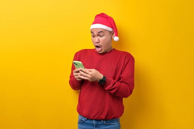 Sorprendido joven asiático con sombrero de Papá Noel usando un teléfono inteligente reaccionando a las noticias en línea sobre la celebración de fondo de estudio amarillo Navidad y concepto de Año Nuevo
