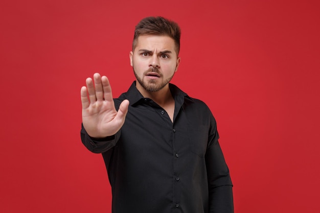 Foto sorprendido joven barbudo de 20 años con camisa negra clásica posando aislado en un retrato de estudio de fondo rojo. gente emociones sinceras concepto de estilo de vida. simulacros de espacio de copia. mostrando gesto de parada con la palma.