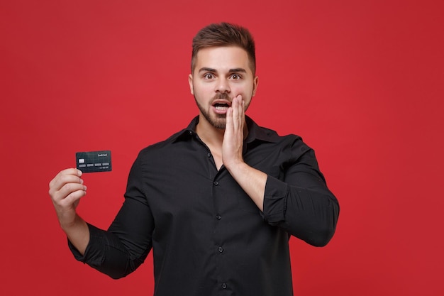 Sorprendido joven barbudo con camisa negra clásica posando aislado en un retrato de estudio de fondo de pared rojo brillante. Concepto de estilo de vida de las personas. Simulacros de espacio de copia. Sostenga la tarjeta de crédito, ponga la mano en la mejilla.