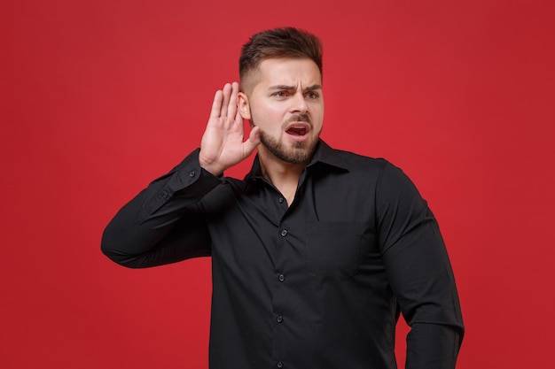 Sorprendido joven barbudo con camisa negra clásica posando aislado en un retrato de estudio de fondo rojo. Emociones sinceras de la gente, concepto de estilo de vida. Simulacros de espacio de copia. Trate de escucharlo con la mano cerca del oído.