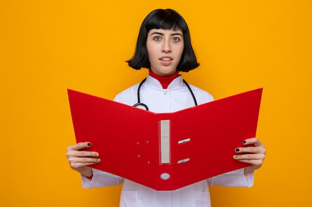 Sorprendido joven bastante caucásica en uniforme médico con estetoscopio sosteniendo la carpeta de archivos y