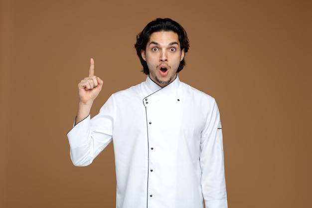 sorprendido joven chef masculino vistiendo uniforme mirando a la cámara apuntando hacia arriba aislado sobre fondo marrón