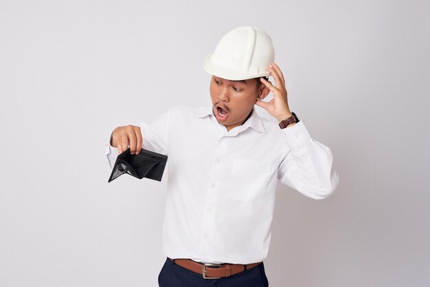 Foto sorprendido joven empleado asiático ingeniero profesional con un casco protector casco mirando la billetera vacía con problemas financieros aislados sobre fondo blanco